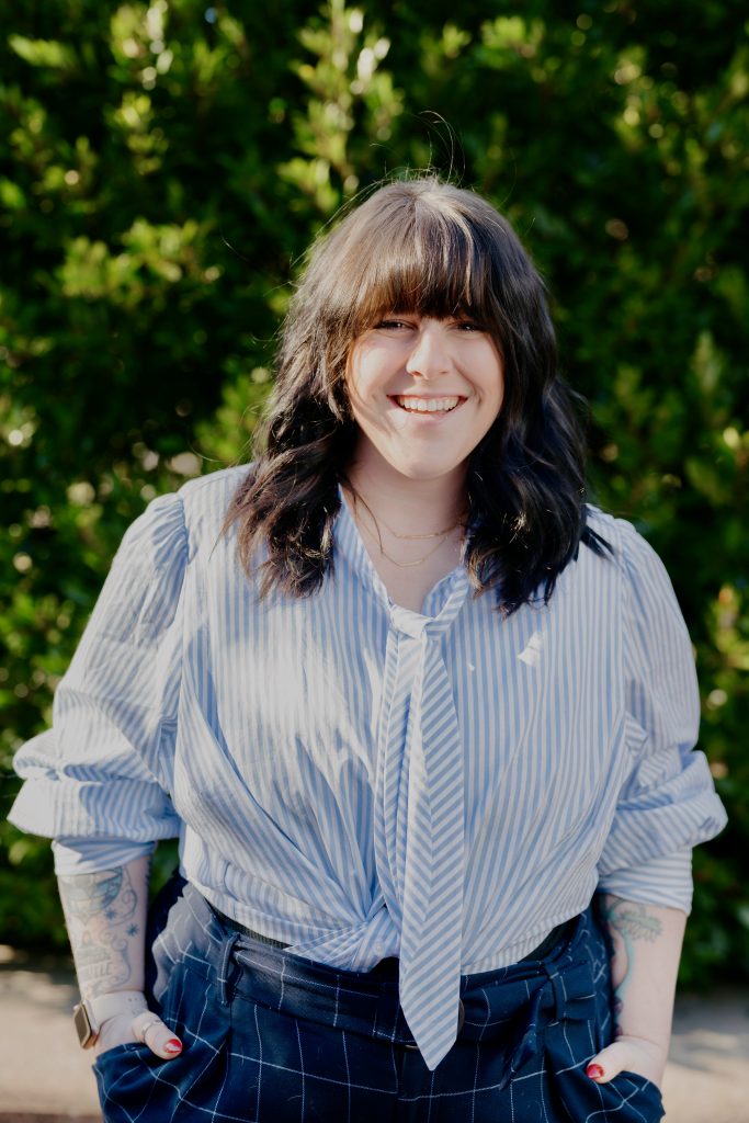 Ashe Anthony, Agent Experience Manager Compass Real Estate= Woman with tattoos, posing in front of a bush.